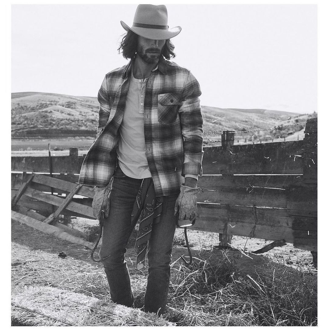 Monochrome portrait of a man in a cowboy hat and plaid shirt holding a lasso on a rural hillside with a wooden fence.
