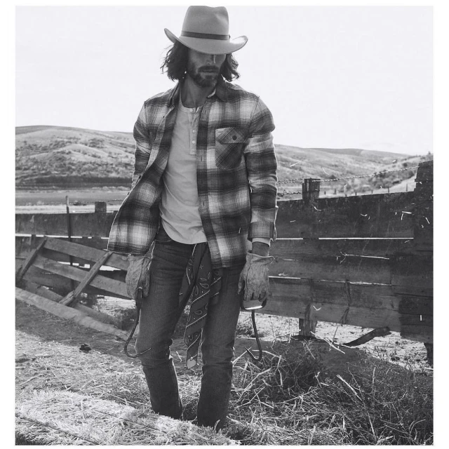Monochrome portrait of a man in a cowboy hat and plaid shirt holding a lasso on a rural hillside with a wooden fence.