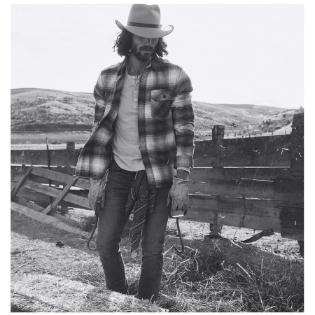 Monochrome portrait of a man in a cowboy hat and plaid shirt holding a lasso on a rural hillside with a wooden fence.