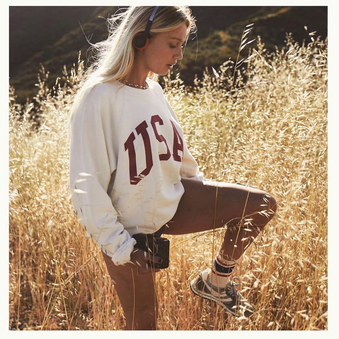 Blond woman in grassy field wearing headphones and USA sweatshirt, holding black digital camera in a natural outdoor setting.