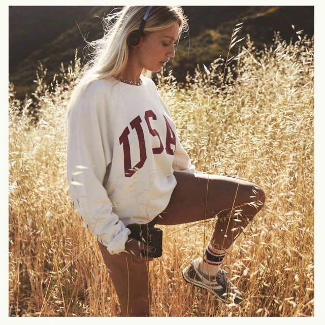 Blond woman in grassy field wearing headphones and USA sweatshirt, holding black digital camera in a natural outdoor setting.