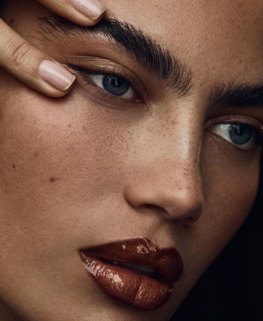 Close-up portrait of a woman with blue eyes and dark hair. She has clear skin with freckles. Her hand is raised to her forehead.
