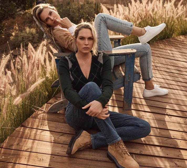 Two adult women pose on a wooden deck with tall grass in the background. One woman sits in a chair, and the other is seated on the deck.