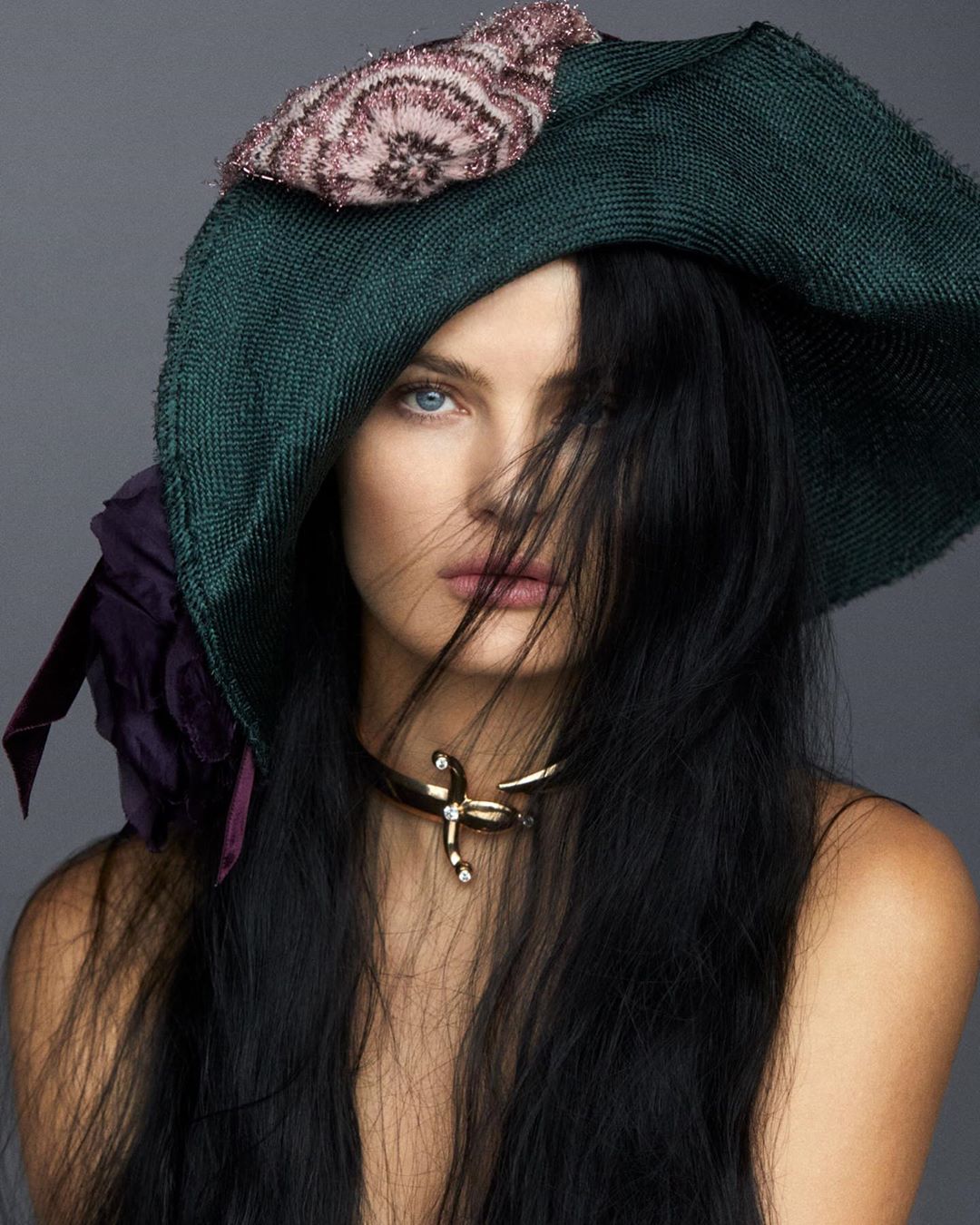 Woman with dark hair wearing an ornate dark green hat with floral accents and a gold-colored necklace, posing in a studio setting.