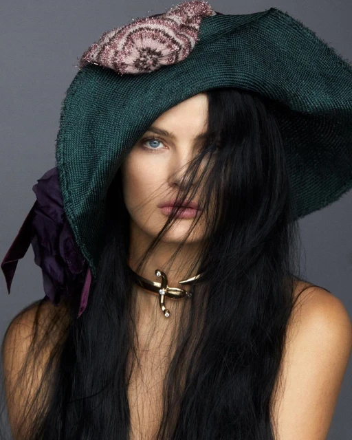 Woman with dark hair wearing an ornate dark green hat with floral accents and a gold-colored necklace, posing in a studio setting.