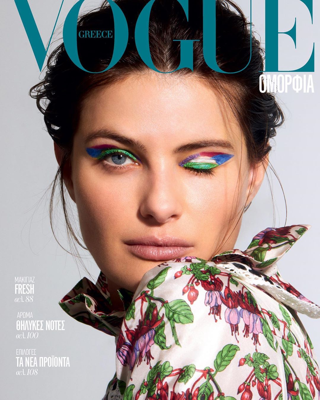 Vogue Greece magazine cover featuring a woman with colorful makeup and a floral top, posing in a studio setting. Beauty and fashion.