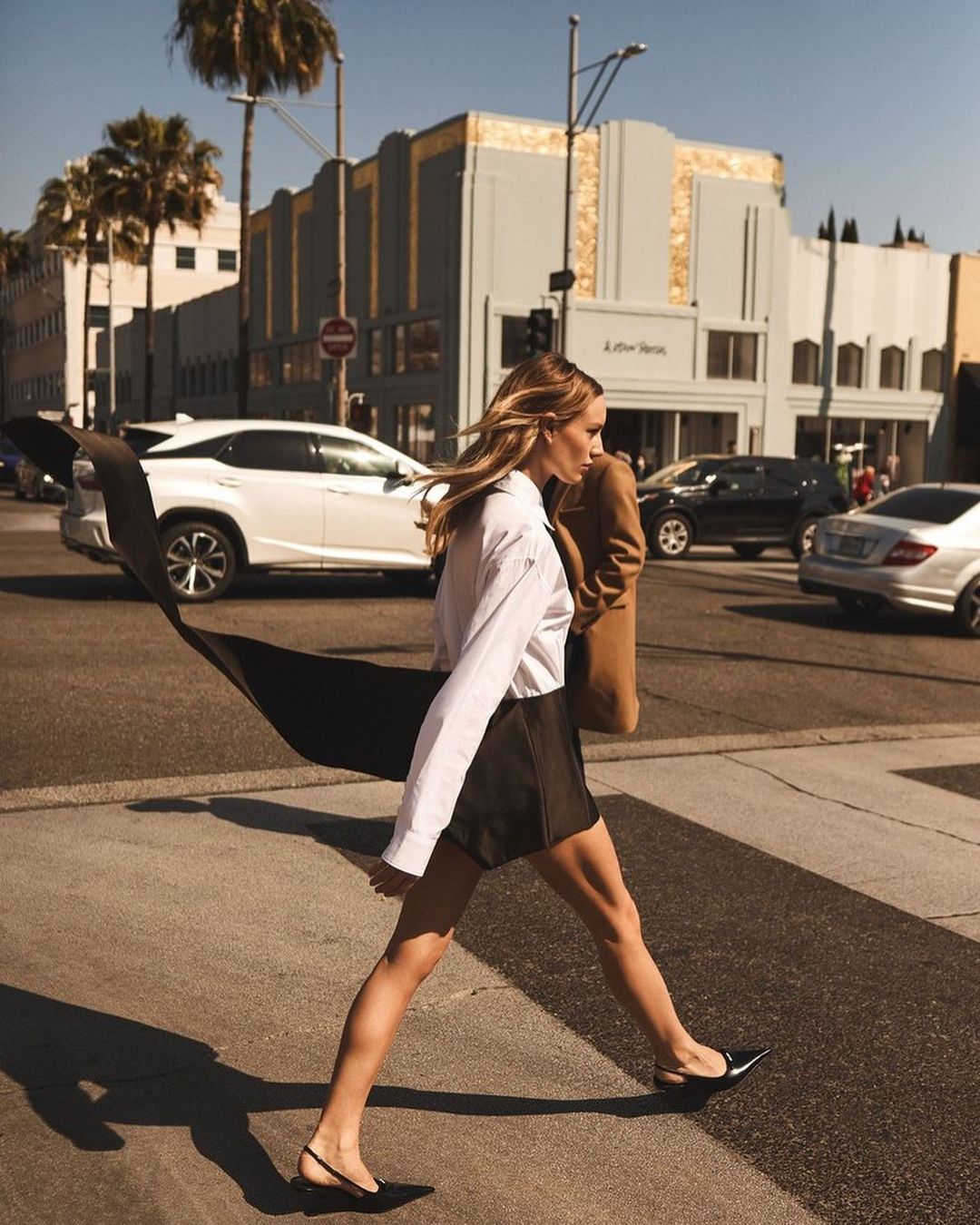 Woman in white shirt and black skirt crosses crosswalk on city street lined with cars and palm trees on a sunny day.