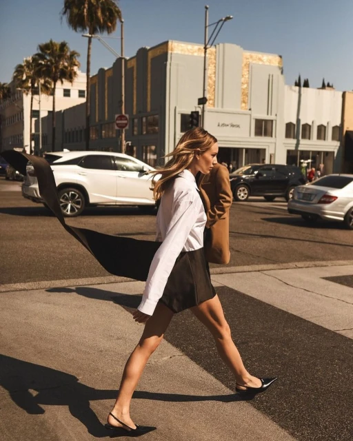 Woman in white shirt and black skirt crosses crosswalk on city street lined with cars and palm trees on a sunny day.