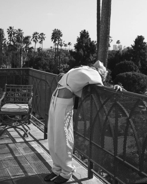 Black and white photo of a woman leaning on a railing, wearing a top and pants, with palm trees and a city in the background.