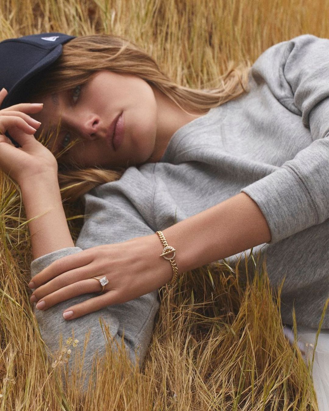 A fair-skinned woman wearing a gray sweatshirt and navy baseball cap lies in a field of golden grass, adorned with a ring and gold bracelet.