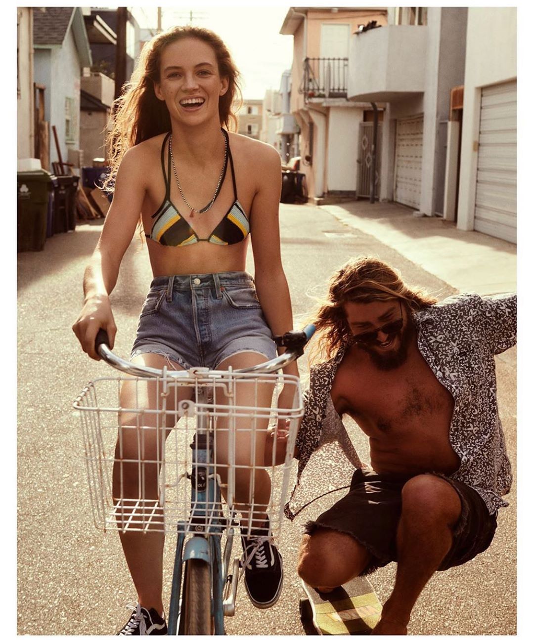 Young woman rides bike in street as man skateboards behind her. Buildings and street fill the background with golden light.