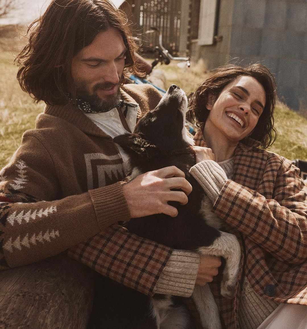 A couple smiles while petting a black and white dog outdoors in the countryside on a sunny day. The woman wears a brown plaid coat.