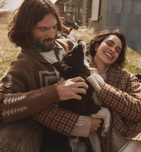 A couple smiles while petting a black and white dog outdoors in the countryside on a sunny day. The woman wears a brown plaid coat.