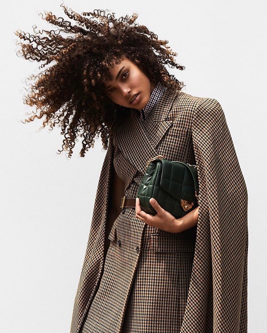 Woman with afro hairstyle poses in a brown and tan houndstooth coat and holds a green quilted leather purse against a plain backdrop.