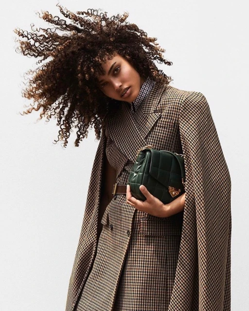 Woman with afro hairstyle poses in a brown and tan houndstooth coat and holds a green quilted leather purse against a plain backdrop.