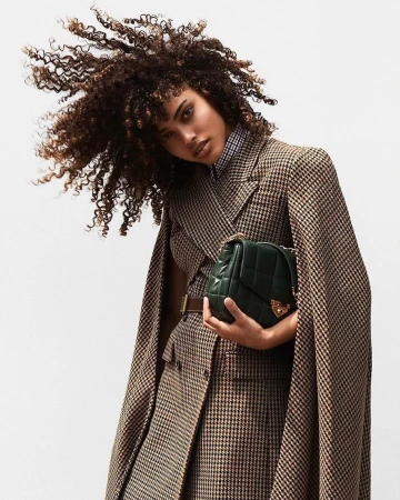 Woman with afro hairstyle poses in a brown and tan houndstooth coat and holds a green quilted leather purse against a plain backdrop.