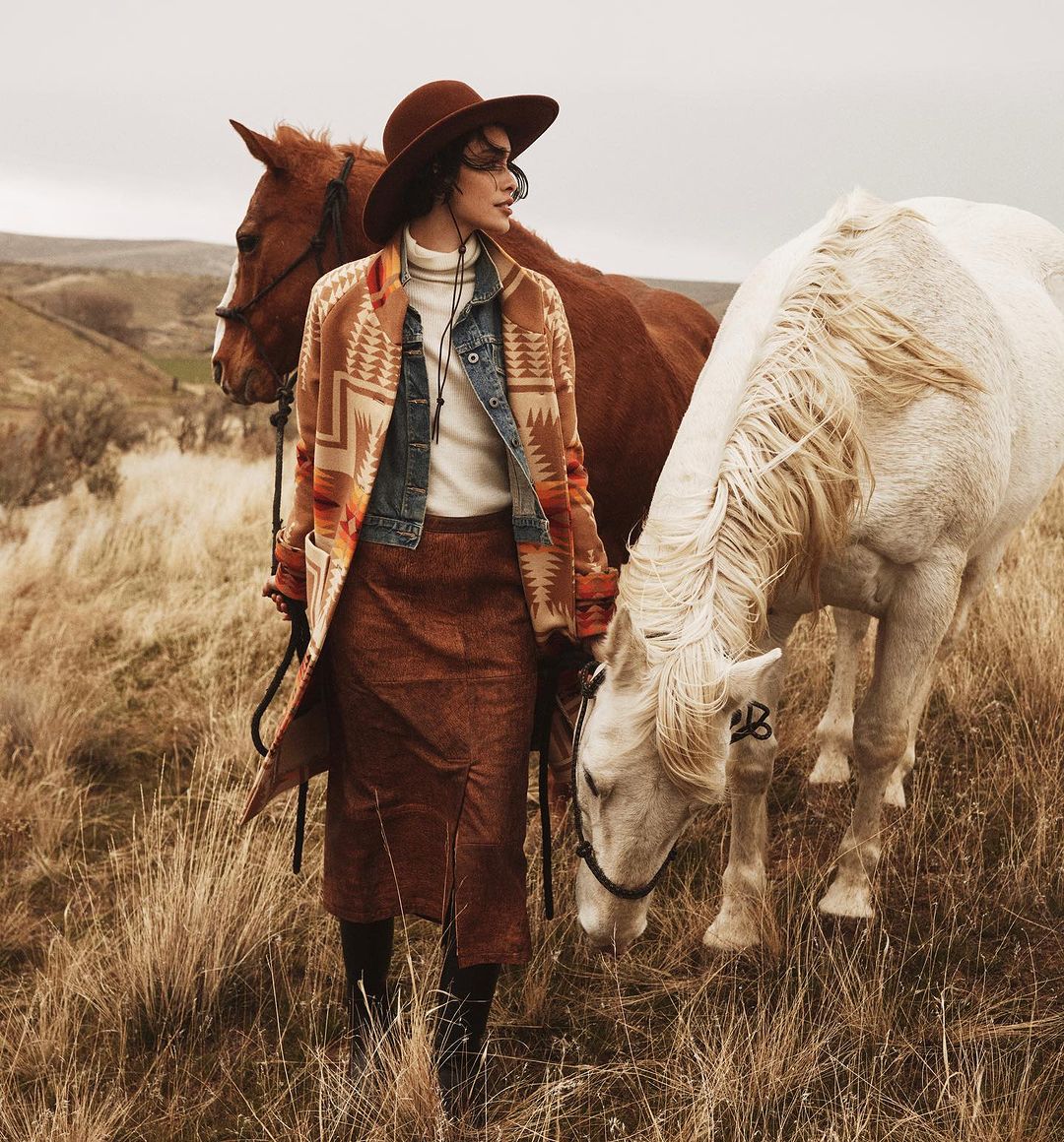 Woman in western-style jacket with horses in rural landscape with beige grasses and overcast skies wearing a brown hat and skirt.