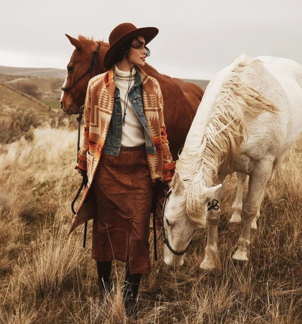 Woman in western-style jacket with horses in rural landscape with beige grasses and overcast skies wearing a brown hat and skirt.