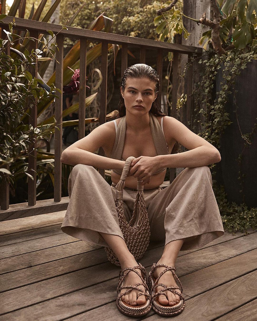 A woman sits cross-legged on a wood deck, wearing a neutral-toned outfit with platform sandals, holding a woven bag, with lush greenery behind.