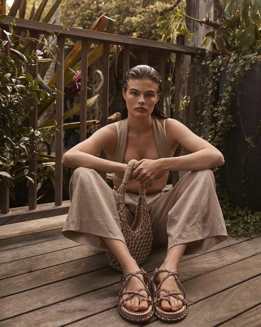 A woman sits cross-legged on a wood deck, wearing a neutral-toned outfit with platform sandals, holding a woven bag, with lush greenery behind.