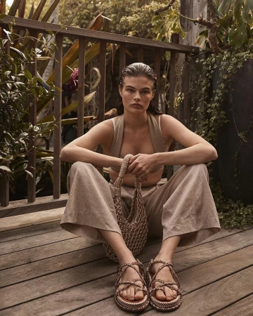 A woman sits cross-legged on a wood deck, wearing a neutral-toned outfit with platform sandals, holding a woven bag, with lush greenery behind.