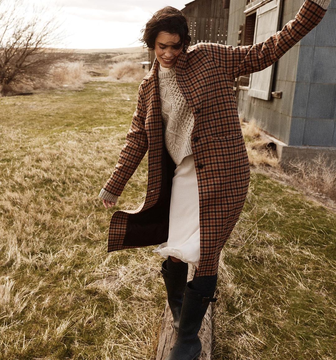 Woman walks on a log in a field wearing a plaid coat, sweater, skirt, and boots. An old barn and dry vegetation are in the background.