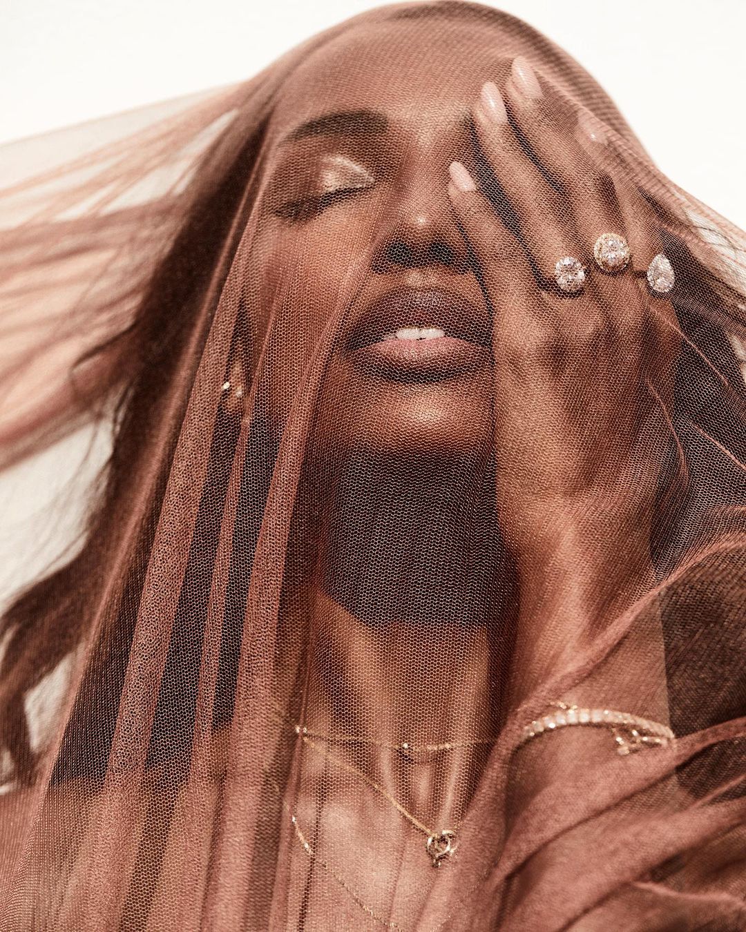 Close-up studio portrait of an adult woman partially veiled, wearing diamond jewelry, neutral makeup, against a white background.