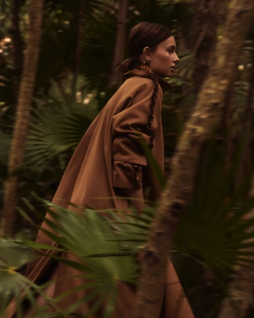 An adult woman walks through the forest, wearing a brown wool coat. The sun shines through the palm trees.