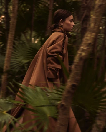 An adult woman walks through the forest, wearing a brown wool coat. The sun shines through the palm trees.