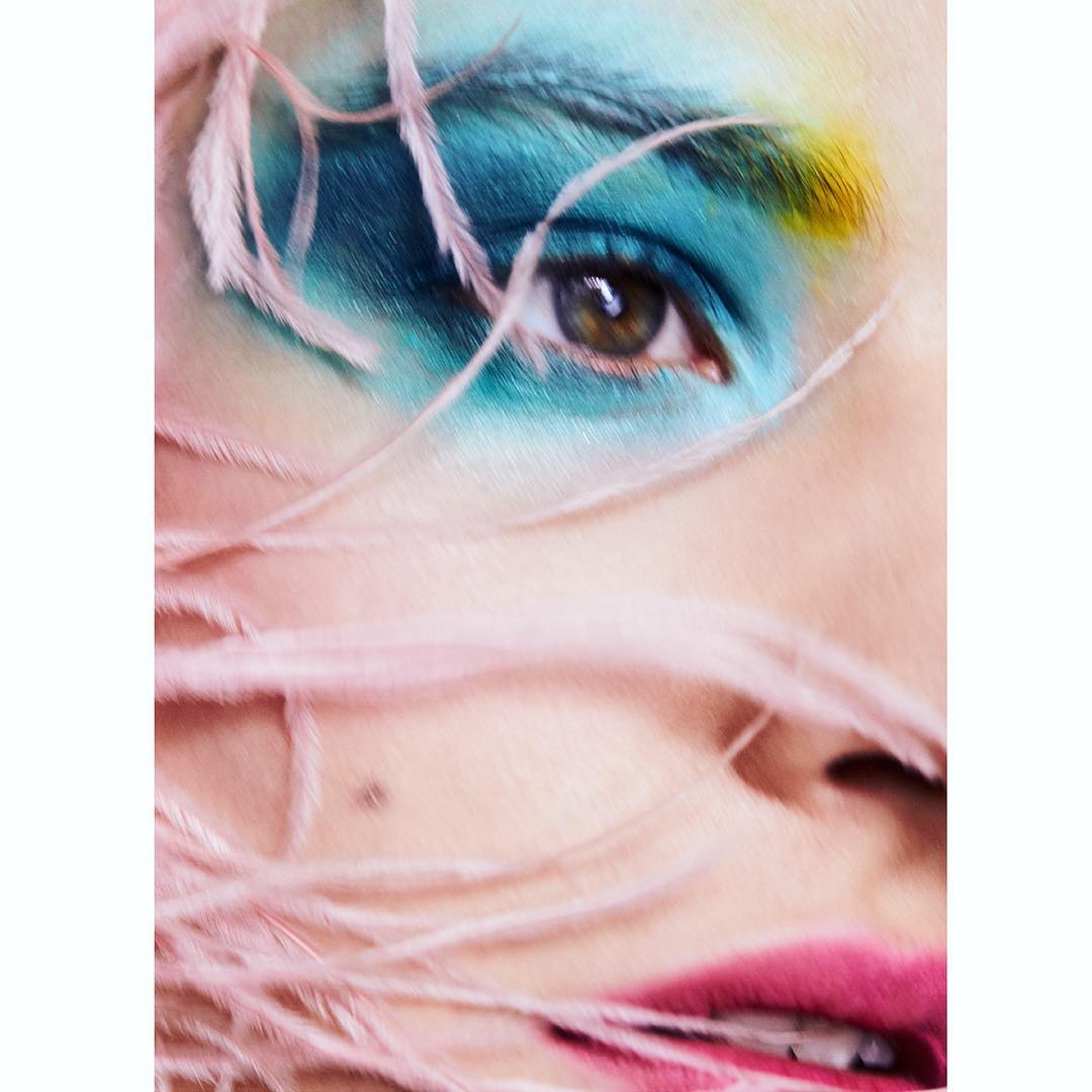 Close-up of a woman's face with blue and yellow eyeshadow and pink lipstick, decorated with soft pink feathers.