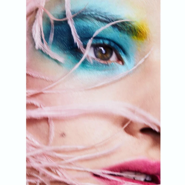 Close-up of a woman's face with blue and yellow eyeshadow and pink lipstick, decorated with soft pink feathers.
