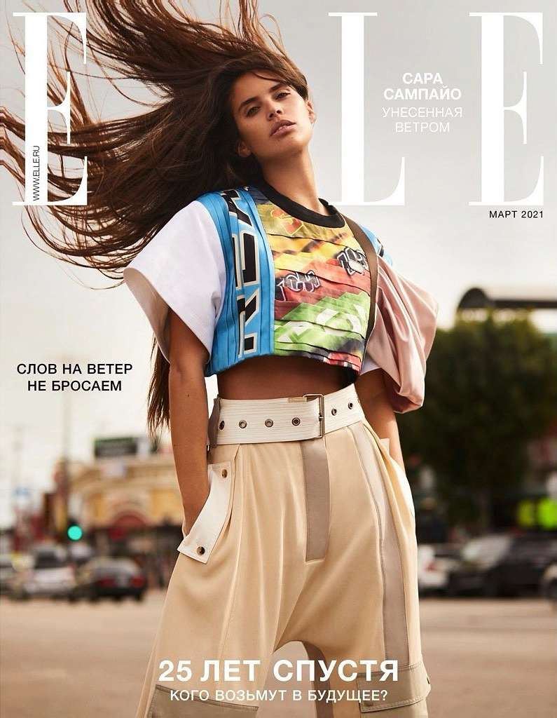 Magazine cover featuring a female model with long brown hair wearing a colorful top and beige pants, posing in an urban environment.