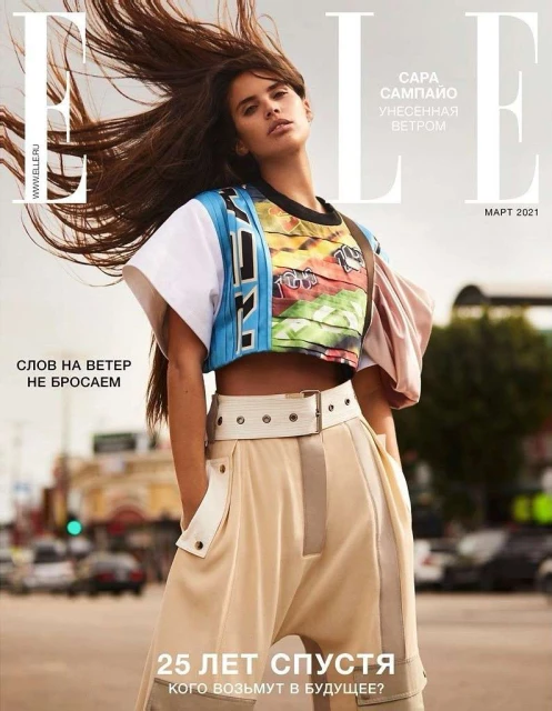 Magazine cover featuring a female model with long brown hair wearing a colorful top and beige pants, posing in an urban environment.