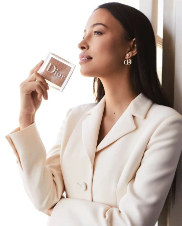 Elegant woman in white blazer holding Dior makeup compact, highlighting beauty and fashion in a clean, bright setting.