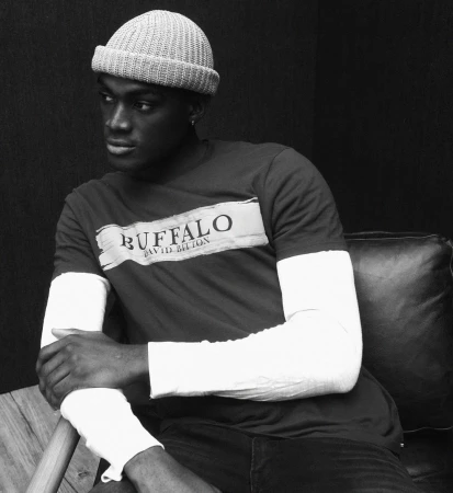 Black man wearing a knit beanie and layered graphic t-shirt seated indoors, photographed by Adam Franzino.