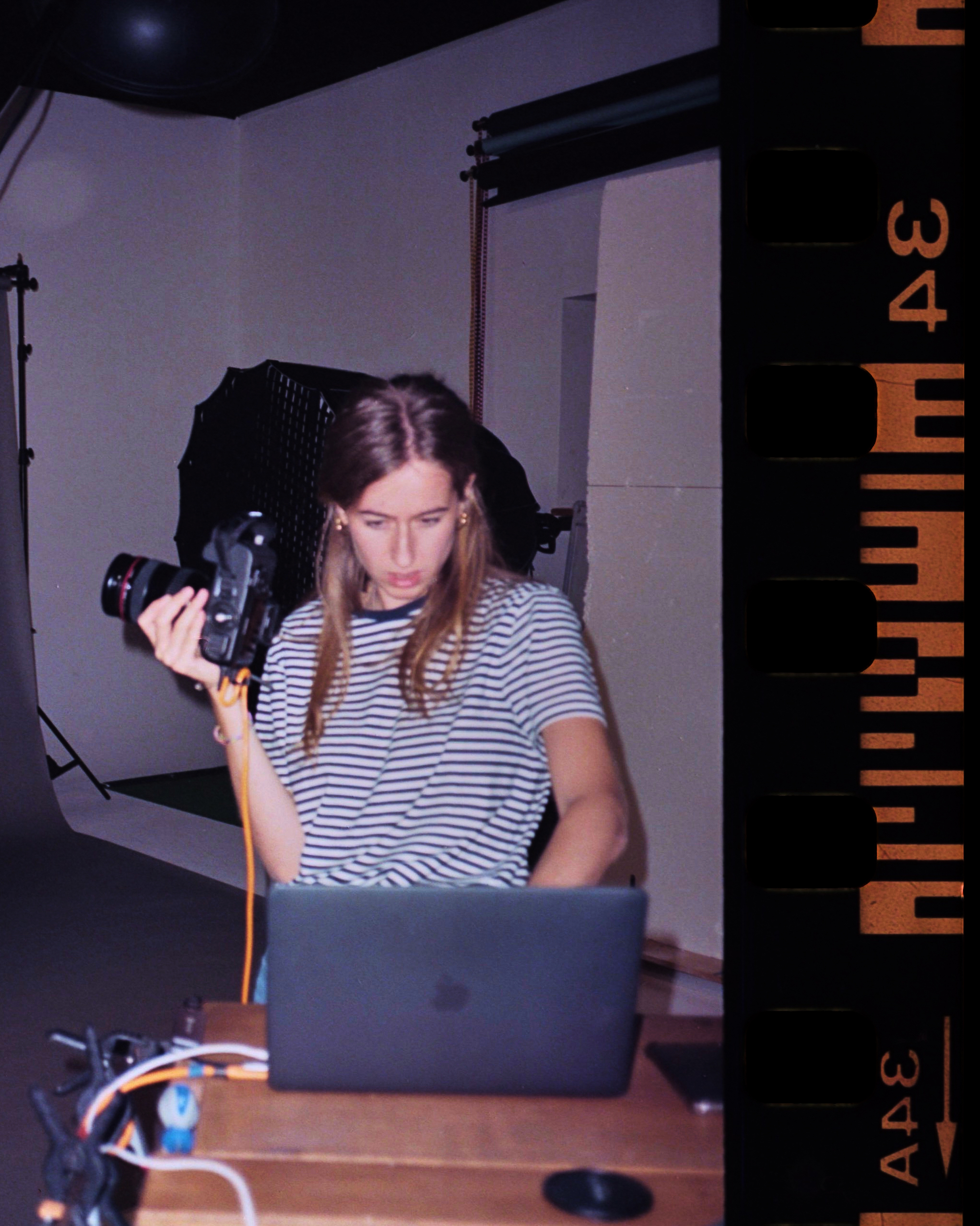 A woman with long hair is holding a digital camera in a photo studio while working on a laptop, with studio lighting and filmstrip border.