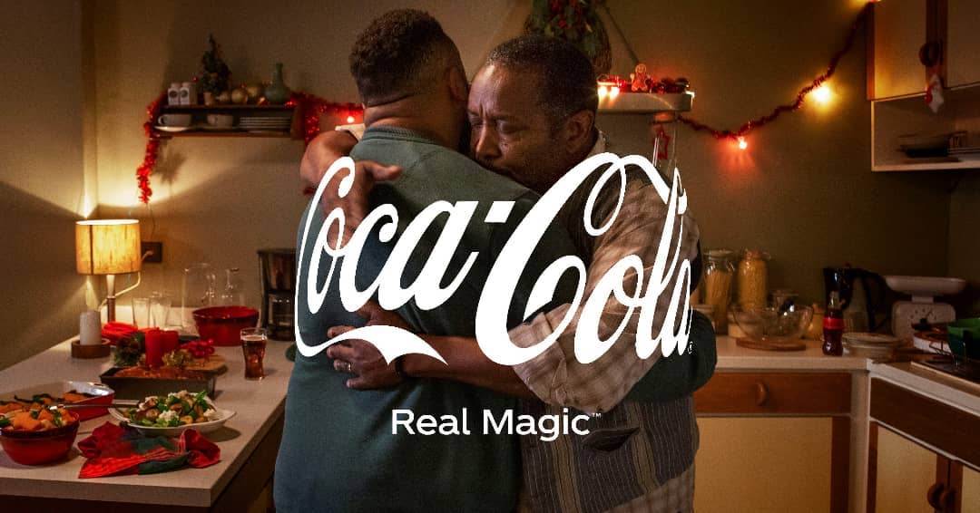 Two men share a heartfelt embrace in a warm kitchen, adorned with holiday decorations, a table laden with food, and the prominent Coca-Cola logo.