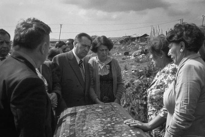 Black and white photo of a group of people gathered outdoors, in what appears to be a rural area, around a decorated object.