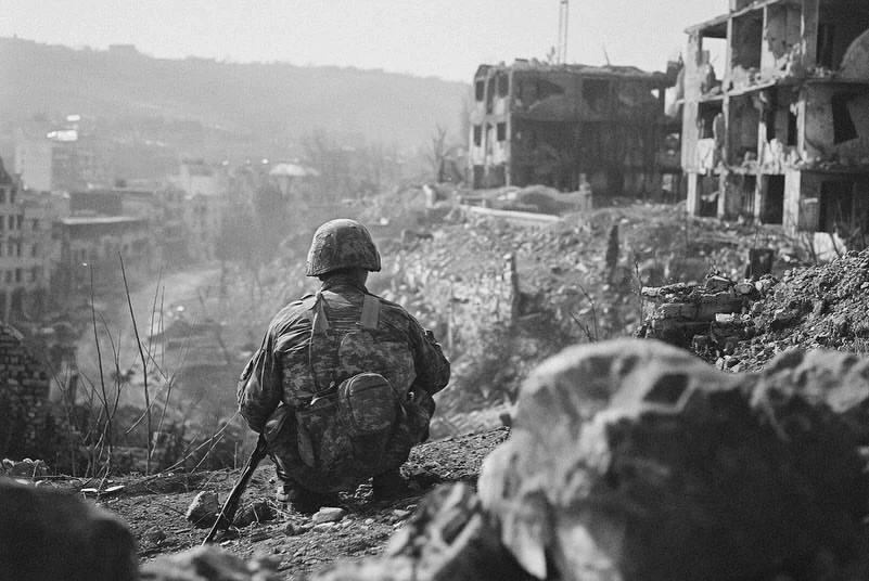 Monochrome image featuring a soldier in camouflage gear crouching, overlooking a war-torn urban landscape with destroyed buildings and rubble.