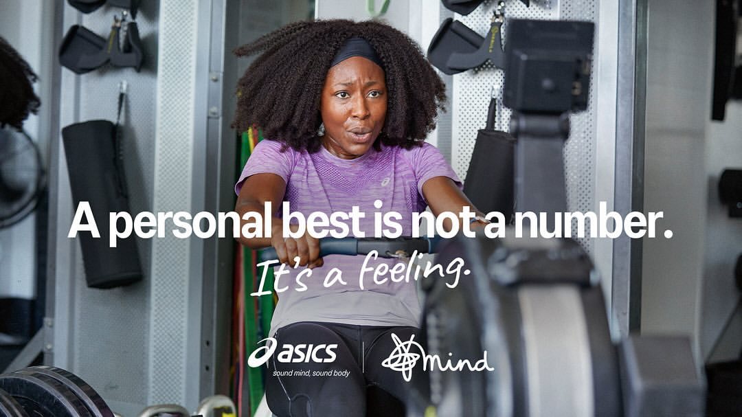 Woman uses rowing machine in gym, determination etched on her face. ASICS and Mind logos visible. Workout promotes mind and body wellness.