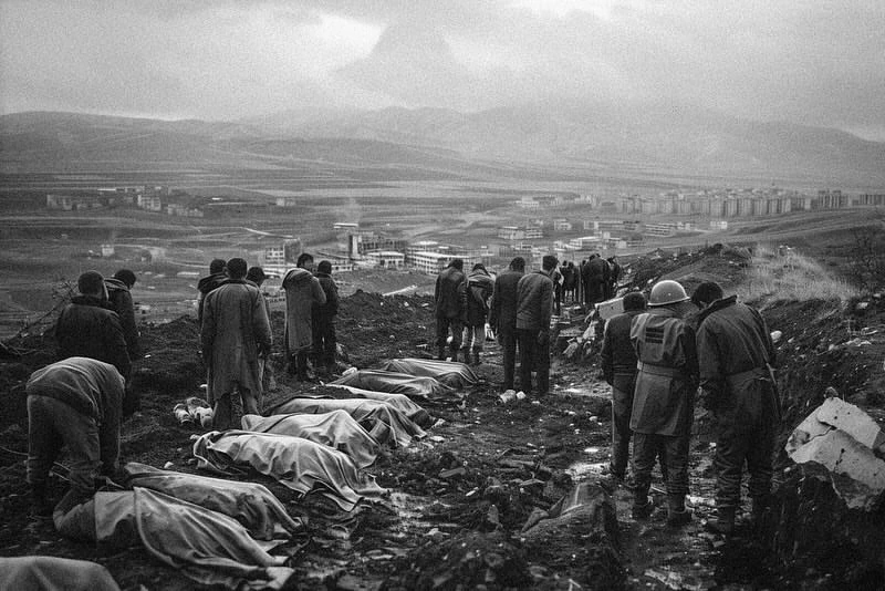 Black and white photograph shows a somber scene with people standing over bodies covered in sheets, against a backdrop of distant buildings and mountains.