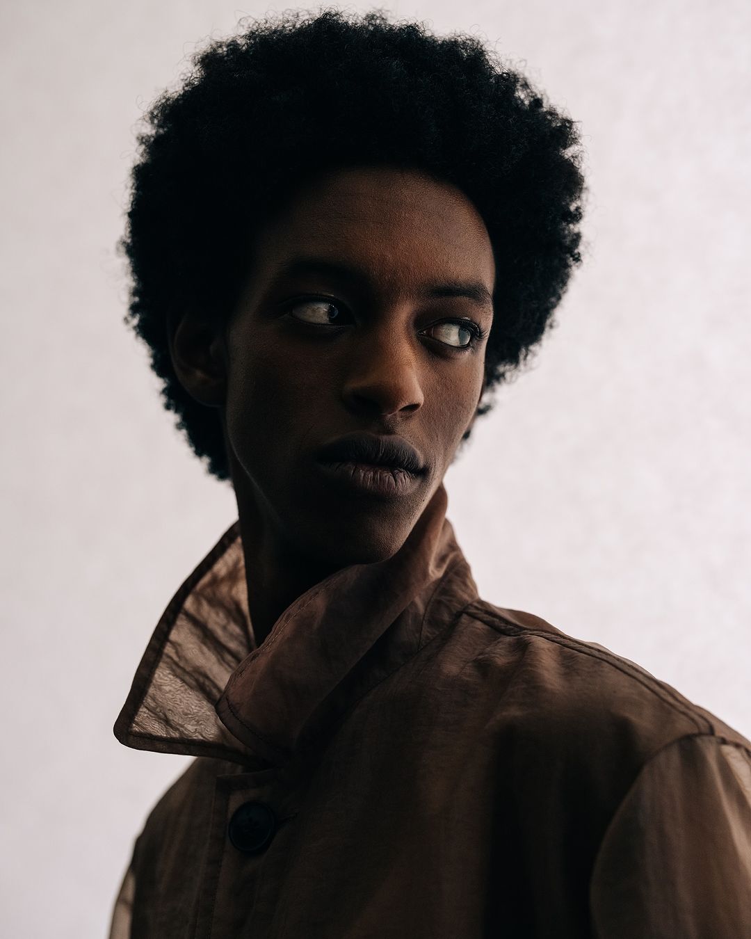Close-up portrait of a young adult with dark skin and curly black hair, wearing a brown shirt with a wide collar, against a plain background.