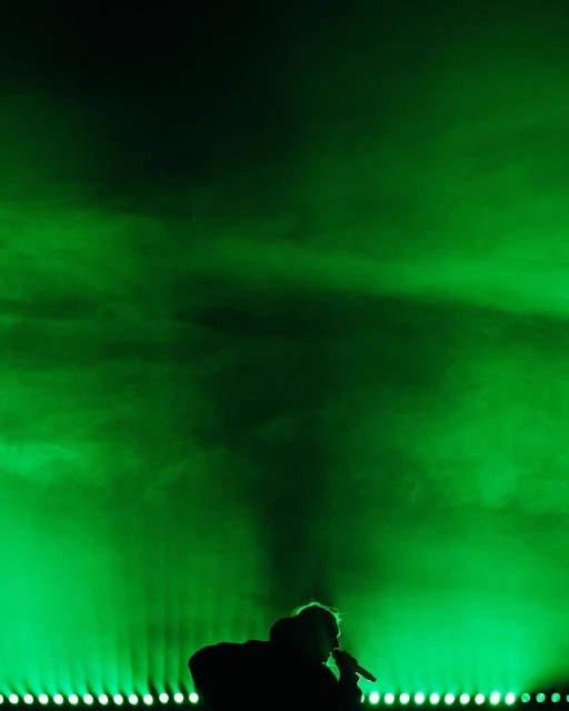Silhouette of a singer performing on stage with a microphone against a vibrant green background with bright lights. Dark figure singing.