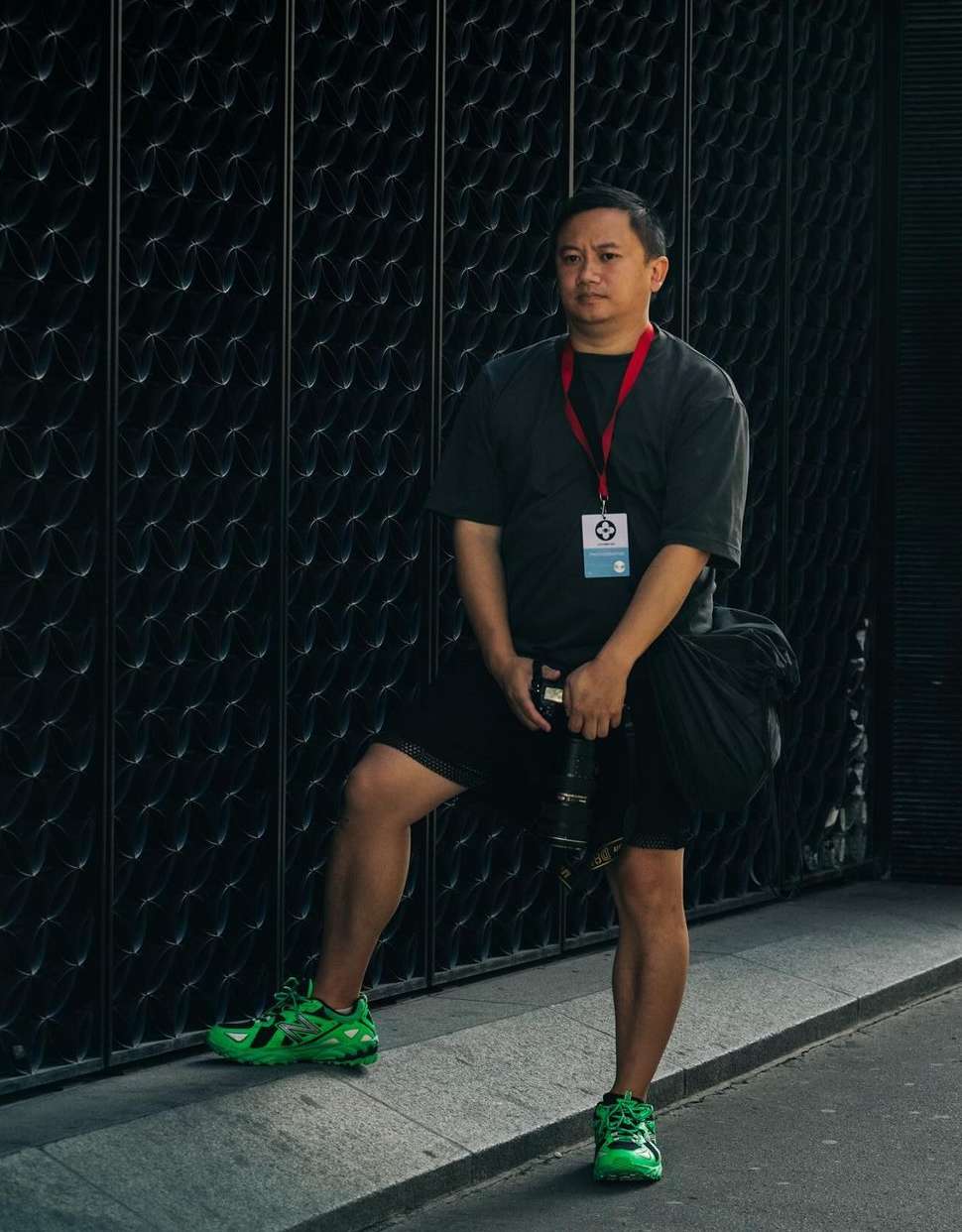 Man stands in front of black textured metal gate wearing a black shirt, black shorts, neon-green tennis shoes, holding a black camera bag.