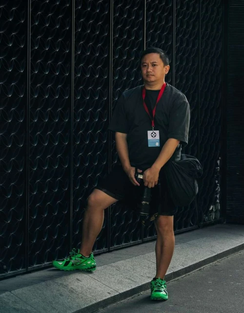 Man stands in front of black textured metal gate wearing a black shirt, black shorts, neon-green tennis shoes, holding a black camera bag.