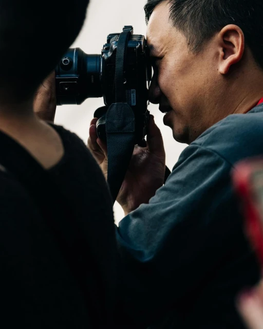 Adult male photographer with professional camera taking a picture. Shoulder of a second person also appears in the photo.