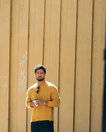Man wearing a mustard yellow knit sweater holding a paper coffee cup against a vertical wooden panel wall.