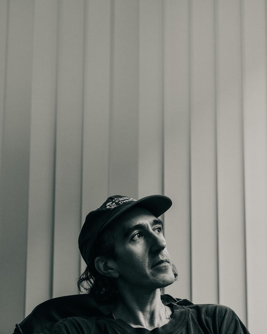Black and white portrait of an adult man wearing a baseball cap looking up in an indoor setting with vertical blinds.