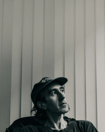Black and white portrait of an adult man wearing a baseball cap looking up in an indoor setting with vertical blinds.