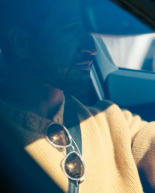 Adult male sits inside car wearing a gold sweater; sunglasses hang from sweater; bright sunlight filters through the windshield.